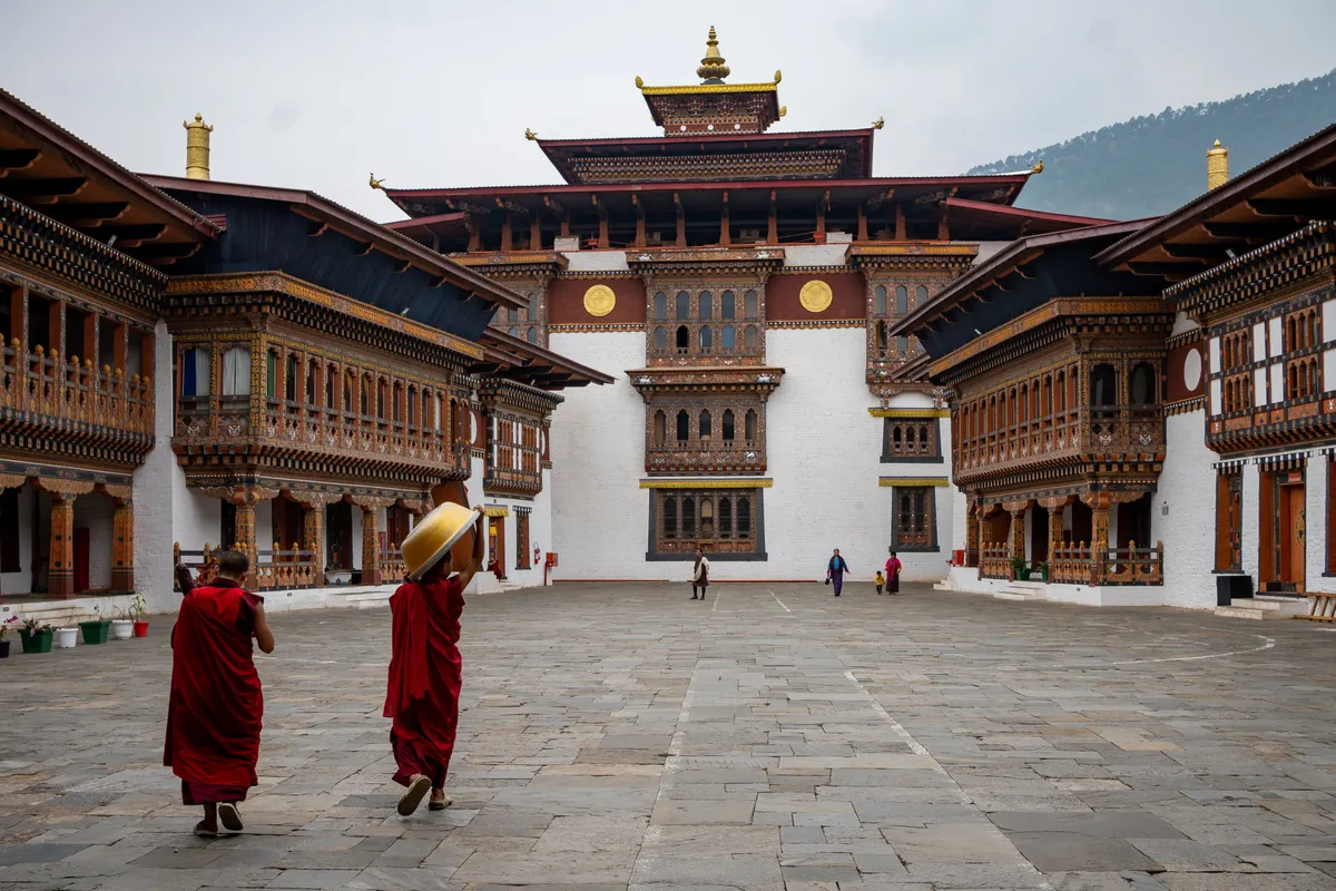 Two robed monks crossing the stone courtyard of Wangdue Phodrang Dzong with ornate painted woodwork, Bhutan