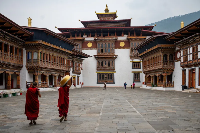 Two robed monks crossing the stone courtyard of Wangdue Phodrang Dzong with ornate painted woodwork, Bhutan