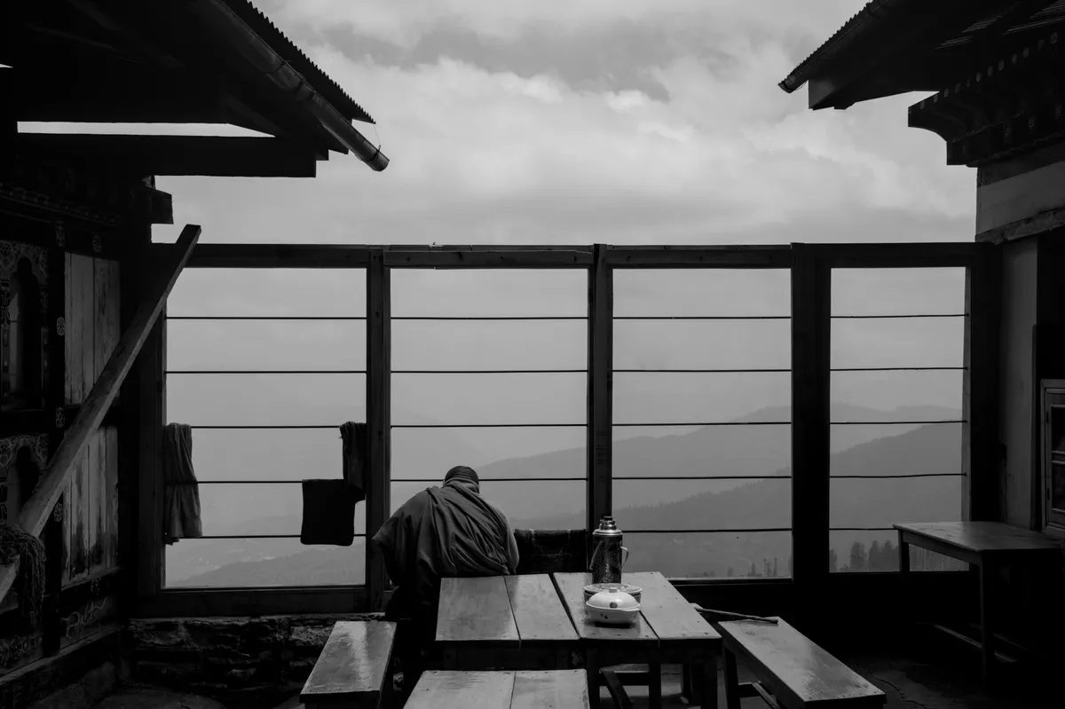 A monk seated at a wooden table on a misty mountain terrace overlooking Himalayan ridges, black and white, Kila Gompa, Bhutan