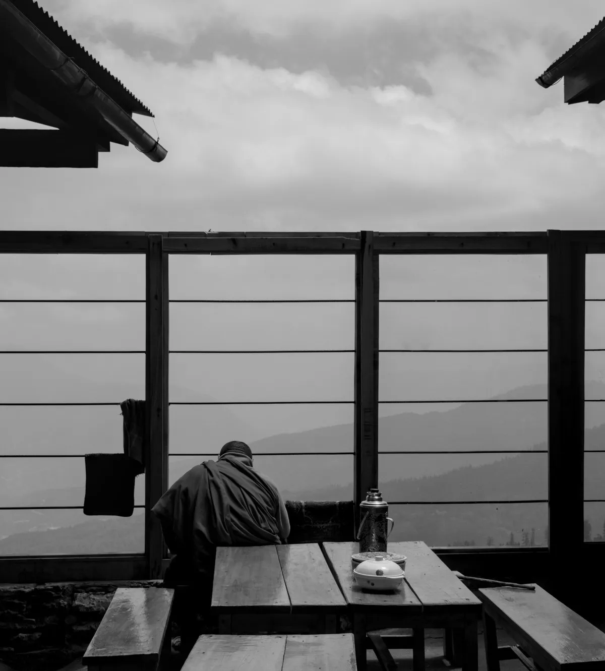 A monk seated at a wooden table on a misty mountain terrace overlooking Himalayan ridges, black and white, Kila Gompa, Bhutan