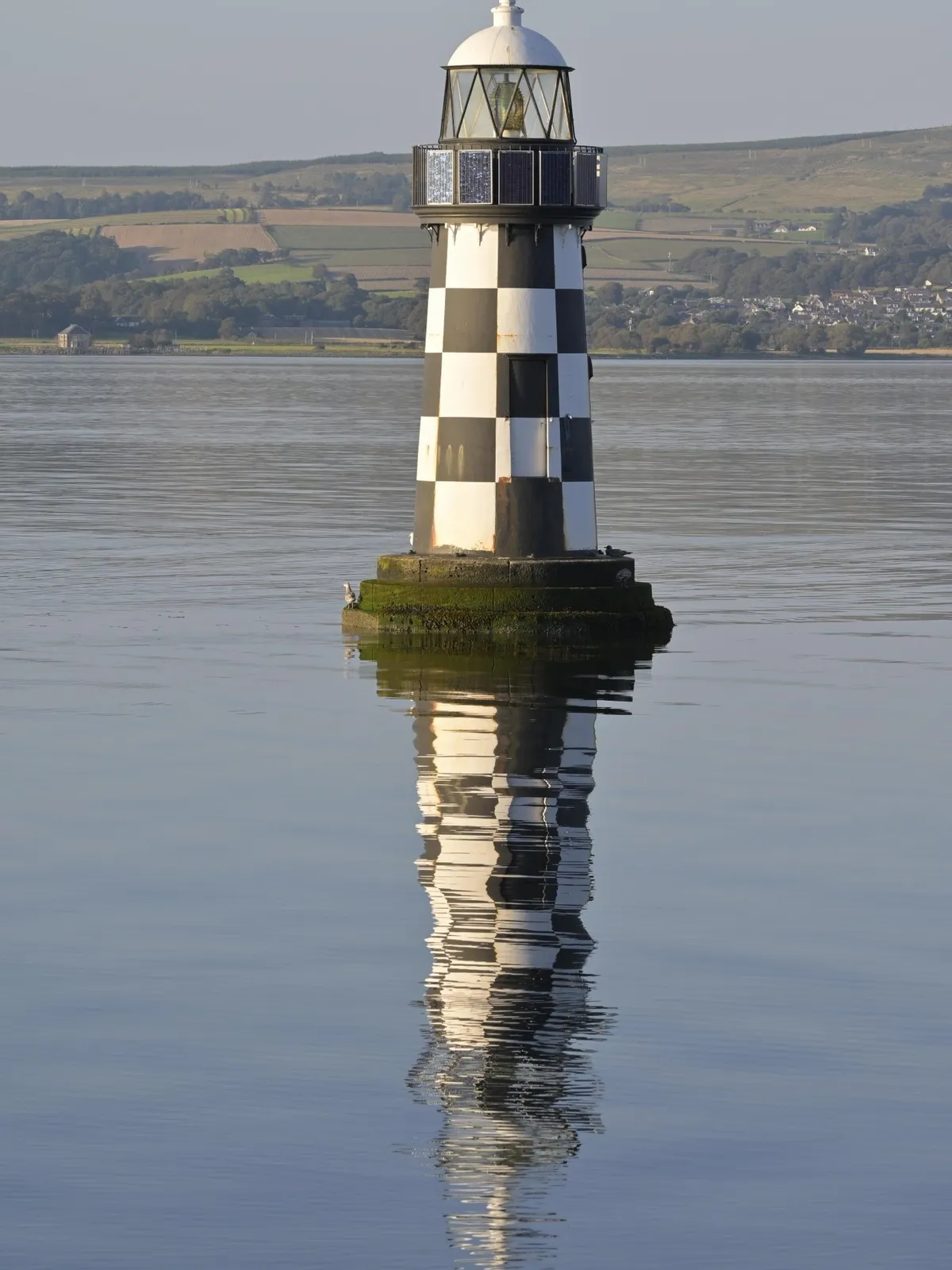 Black and white chequered lighthouse reflected in the calm water of the Firth of Clyde with green hills behind, Scotland