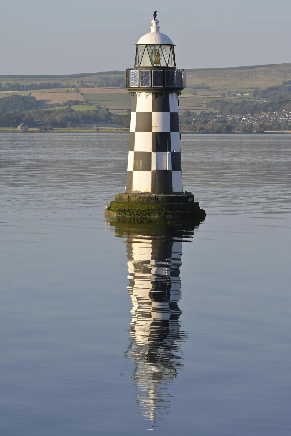 Black and white chequered lighthouse reflected in the calm water of the Firth of Clyde with green hills behind, Scotland