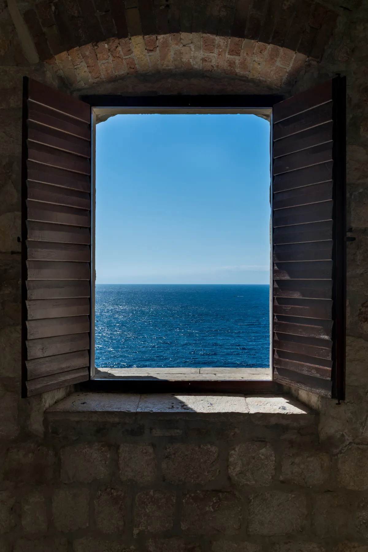 Open wooden-shuttered window in a stone wall framing the deep blue Adriatic sea, Dubrovnik, Croatia