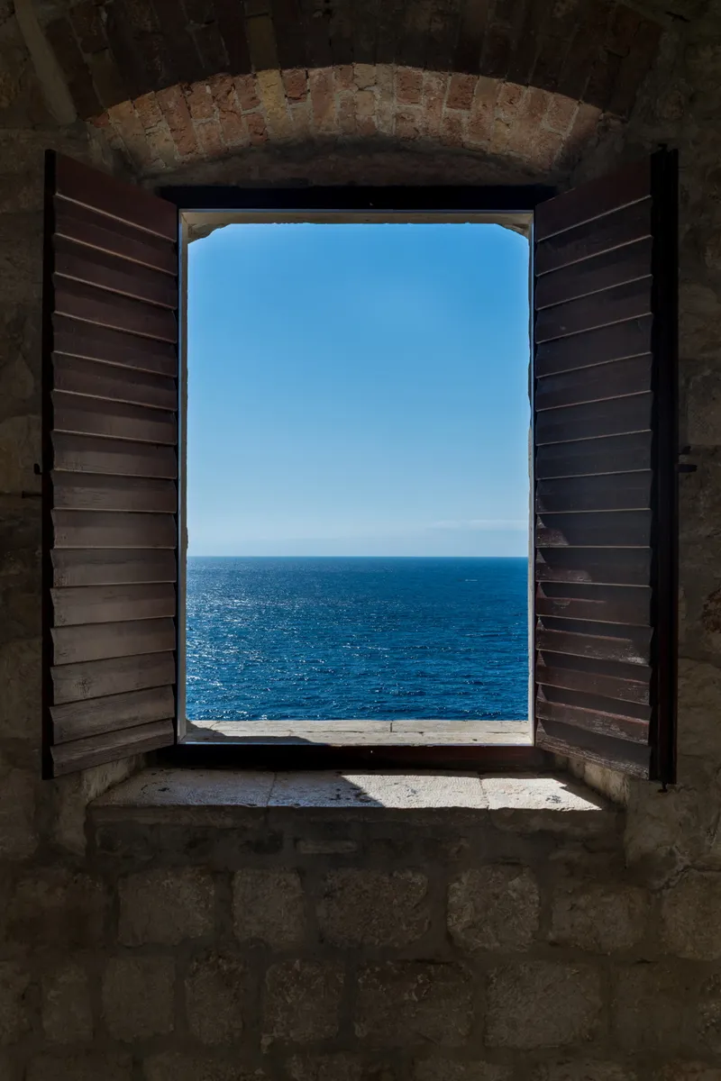 Open wooden-shuttered window in a stone wall framing the deep blue Adriatic sea, Dubrovnik, Croatia