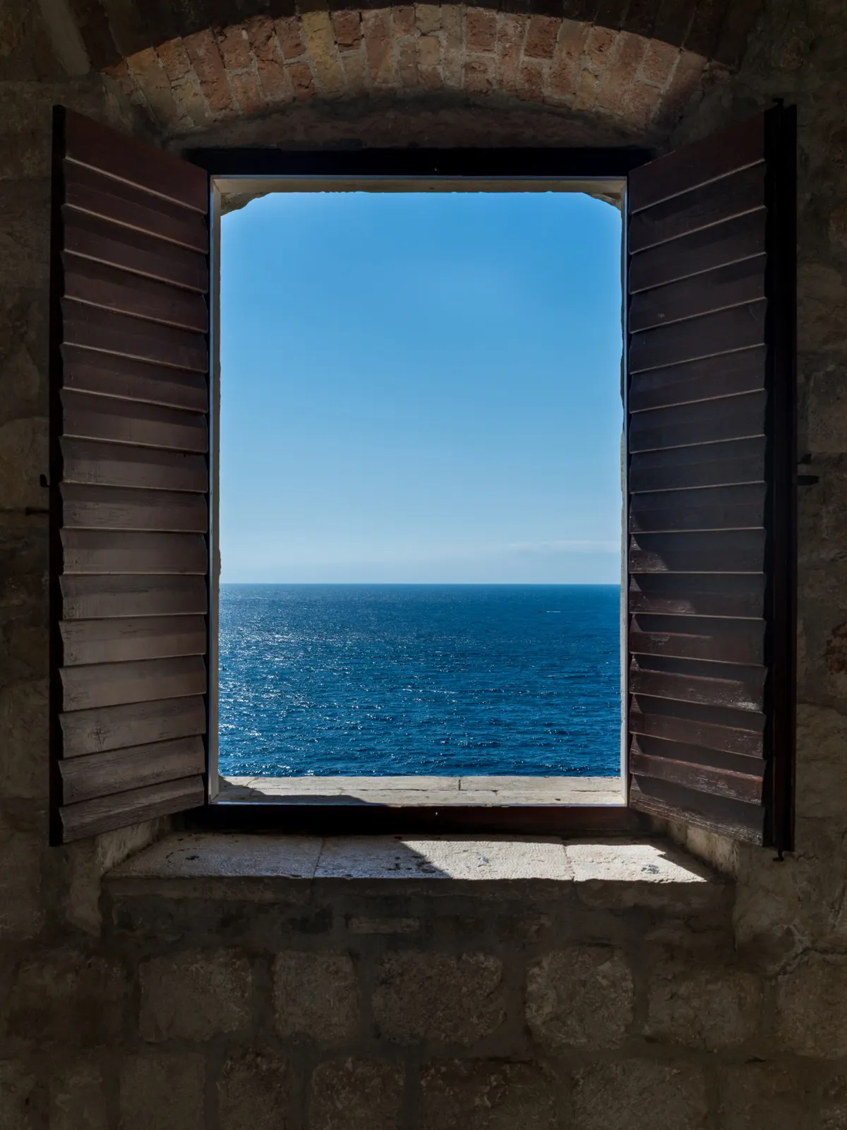Open wooden-shuttered window in a stone wall framing the deep blue Adriatic sea, Dubrovnik, Croatia