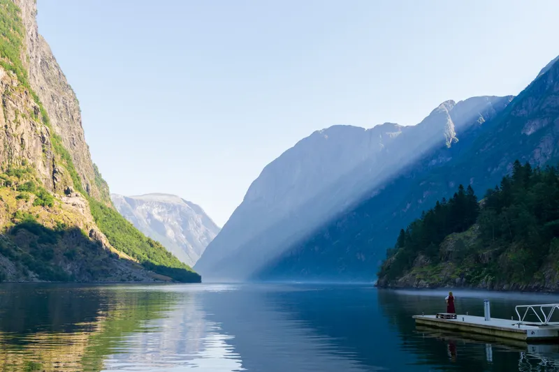 Calm Nærøyfjord with steep mountains, mirror-still water and a lone figure in red on a jetty, Norway