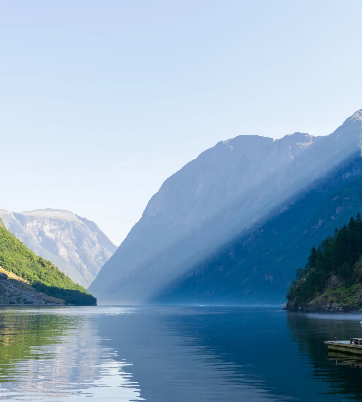 Calm Nærøyfjord with steep mountains, mirror-still water and a lone figure in red on a jetty, Norway