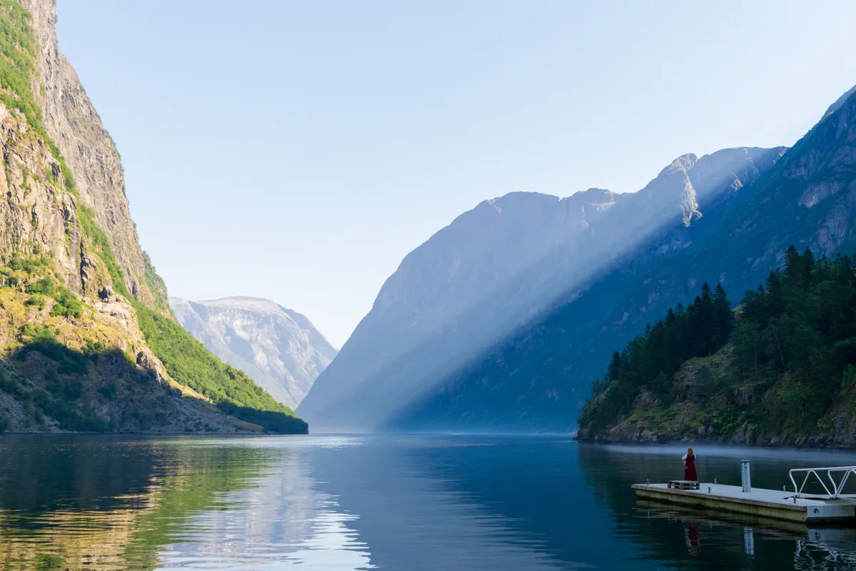 Calm Nærøyfjord with steep mountains, mirror-still water and a lone figure in red on a jetty, Norway